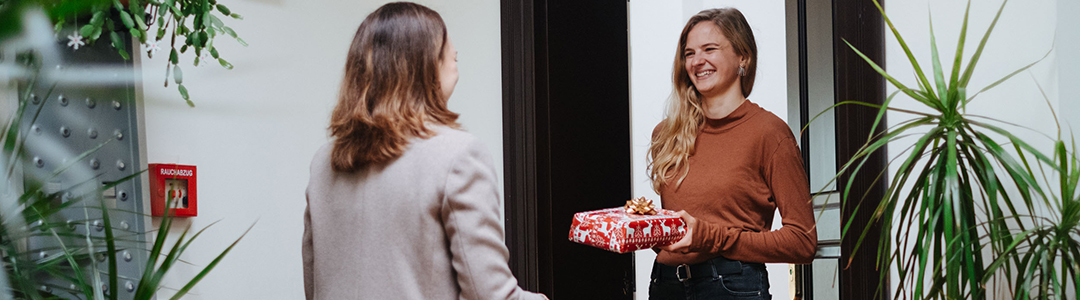 Zwei Frauen stehen sich fröhlich an einer Wohnungstür gegenüber, eine hält ein Geschenk in der Hand